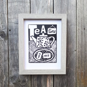 May include: Framed black and white art print with the words "Tea time" and a teapot design. The teapot has a polka dot pattern. The print also includes the word "NICE" twice. The frame is light gray.