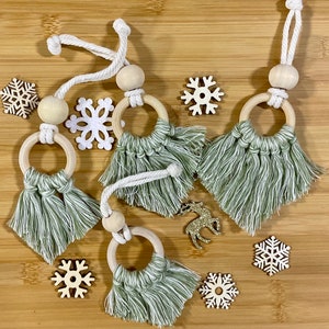 May include: Four handmade macrame ornaments with wooden rings and tassels. The ornaments are in a sage green color and have a white rope. The ornaments are on a wooden surface with white snowflakes.