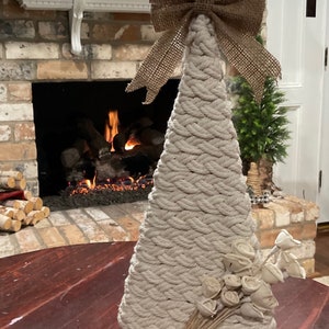 May include: A white rope Christmas tree with a burlap bow on top. The tree is decorated with white wooden roses and is sitting on a wooden base. The tree is in front of a brick fireplace with a fire burning inside.