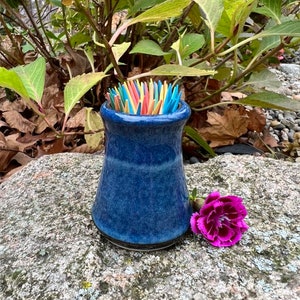 May include: A blue ceramic toothpick holder with a dark blue glaze. The holder is filled with colourful toothpicks and is sitting on a grey rock with a pink flower next to it.