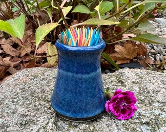 Könnte beinhalten: Ein blauer Keramik-Zahnstocherhalter mit dunkelblauer Glasur. Der Halter ist mit bunten Zahnstochern gefüllt und steht auf einem grauen Stein mit einer rosa Blume daneben.