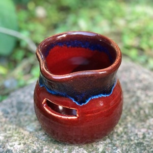 May include: A handmade ceramic oil warmer with a rich, reddish-brown glaze. The rim and upper portion are a darker brown, with a vibrant blue accent. A rectangular opening is visible on the side. The warmer is designed for aromatherapy.