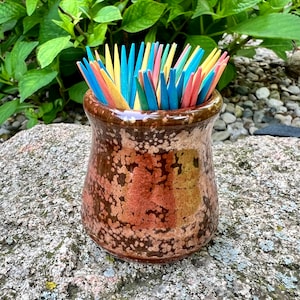 May include: A brown ceramic toothpick holder with a speckled glaze. The holder is filled with colorful toothpicks in shades of yellow, blue, and pink.