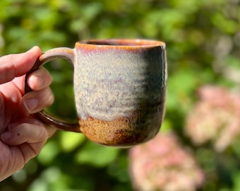 May include: A ceramic mug with a rounded handle, featuring a reactive glaze finish that transitions from a speckled light blue and cream top to a textured, earthy brown base with a dark rim around the top edge of the vessel.