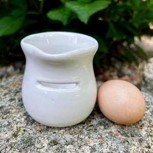 May include: A small, white ceramic pitcher with a curved spout and a horizontal slot, next to a brown egg. The pitcher has a smooth, glossy finish. The egg is oval-shaped and has a speckled appearance. The items are on a textured, gray stone surface.