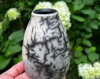 May include: A small, white ceramic vase with a black crackle glaze. The vase has a rounded, egg-shaped body and a narrow opening.