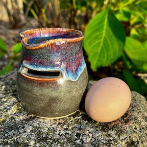 May include: A dark gray ceramic egg holder with a purple and blue glaze. The holder has a small hole for the egg to sit in. A brown egg is sitting next to the holder.