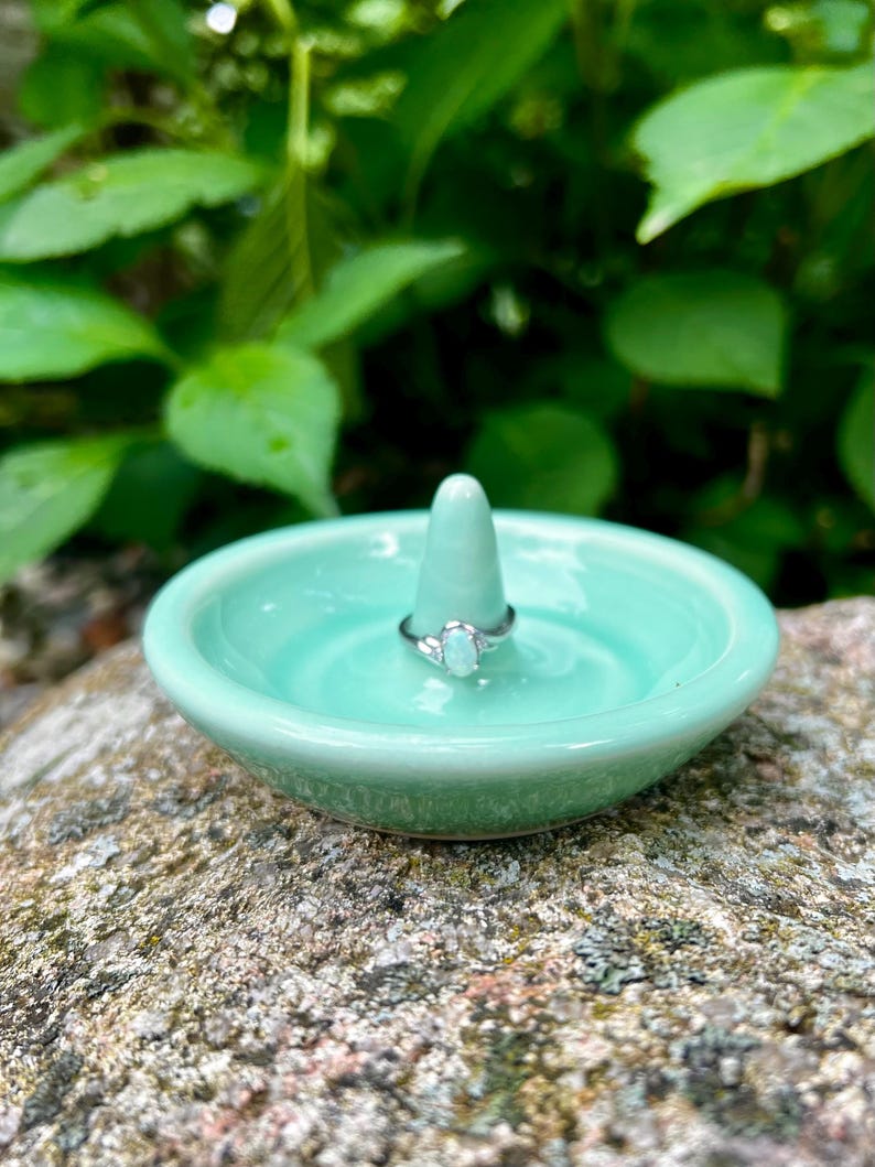 Puede incluir: Un porta anillos de cer&aacute;mica verde claro con un centro en forma de cono. Un anillo de plata con una piedra ovalada blanca descansa sobre el cono.