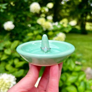 Puede incluir: Un porta anillos de cer&aacute;mica verde claro con un peque&ntilde;o soporte c&oacute;nico para anillos. Un anillo de plata con una piedra ovalada transparente est&aacute; en el soporte.