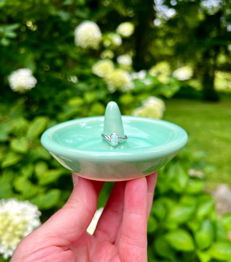 Puede incluir: Un porta anillos de cer&aacute;mica verde claro con un peque&ntilde;o soporte c&oacute;nico para anillos. Un anillo de plata con una piedra ovalada transparente est&aacute; en el soporte.