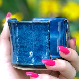 May include: A small, handcrafted blue ceramic cup with a unique, textured surface. The cup features visible gold accents along the edges and a visible crack, showcasing the Kintsugi repair technique. The cup is held in a hand against a blurred background.