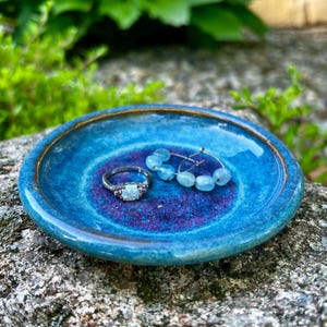 Könnte beinhalten: Eine handgefertigte, runde Keramik-Schmuckschale in Blau- und Lilatönen mit einem bronzefarbenen Rand. Die Schale enthält einen Silberring mit einem Opalstein und ein Paar blaue Perlen-Ohrringe. Die Schale wird auf einer strukturierten Felsfläche präsentiert.