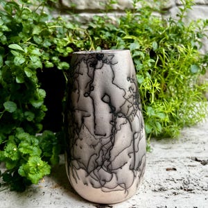 Könnte beinhalten: Eine handgefertigte Keramikvase mit einer einzigartigen, strukturierten Oberfläche. Die Vase hat einen hellgrauen Sockel mit komplizierten schwarzen, blitzartigen Mustern. Die Vase ist zylindrisch mit einem leicht abgerundeten Boden. Der Hintergrund zeigt grüne Pflanzen und eine Steinmauer.