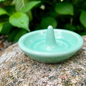 Puede incluir: Un porta anillos de cer&aacute;mica azul claro con un centro elevado para sujetar los anillos. El plato est&aacute; sobre una roca.