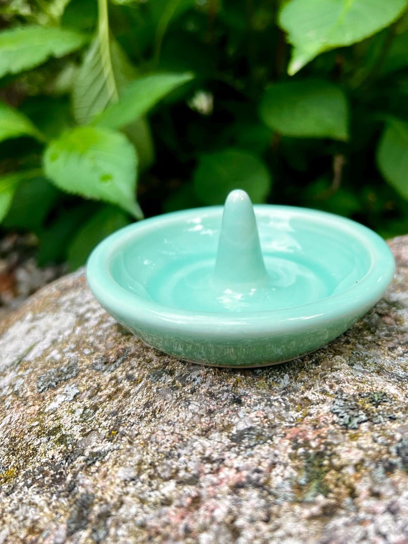 Puede incluir: Un porta anillos de cer&aacute;mica azul claro con un centro elevado para sujetar los anillos. El plato est&aacute; sobre una roca.