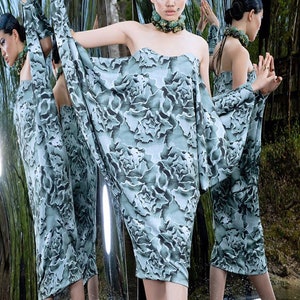 May include: A woman wearing a long, flowing, off-the-shoulder dress with a floral print. The dress is a light blue and green color. The woman is standing in a shallow pool of water, with a mirrored background.