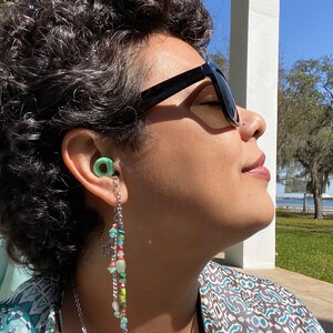 May include: A person wearing sunglasses and a green earplug with a beaded chain hanging from it. The chain has colorful beads, including green, blue, red, and white.