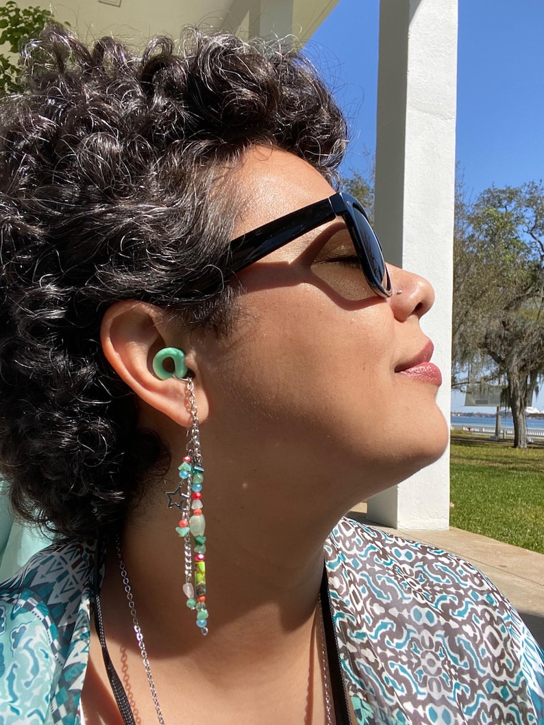 May include: A person wearing sunglasses and a green earplug with a beaded chain hanging from it. The chain has colorful beads, including green, blue, red, and white.