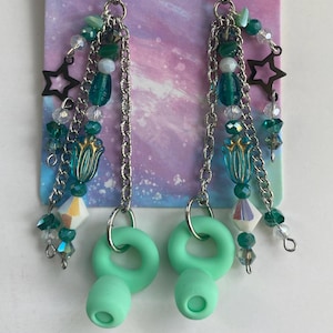 May include: A pair of teal green dangle earrings with a silver chain, beads, and charms. The charms include stars, flowers, and small clear crystals. The earrings are attached to a pair of teal green silicone earplugs.
