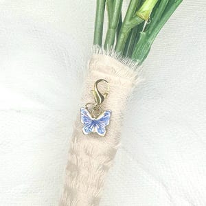 May include: A gold-colored butterfly charm with blue and white enamel detailing, attached to a bouquet of flowers wrapped in beige fabric. The butterfly charm is a delicate accessory.