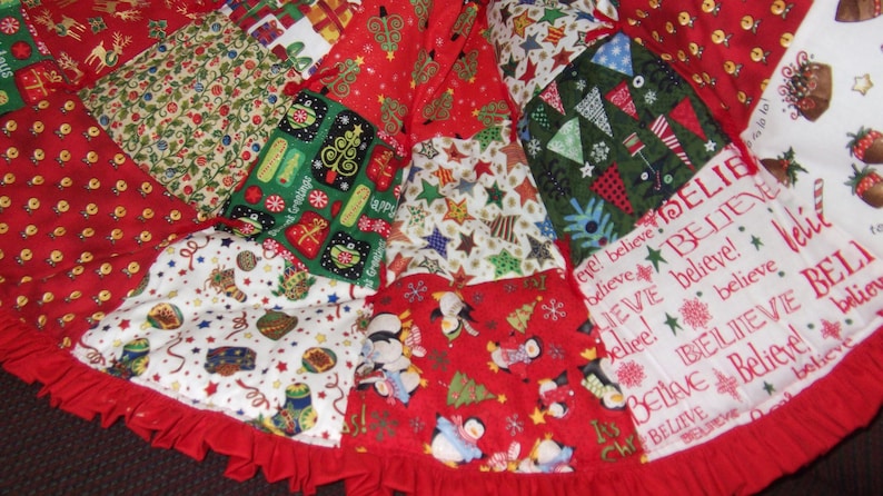 K&ouml;nnte beinhalten: Ein Patchwork-Weihnachtsbaumrock mit rotem R&uuml;schenrand. Der Rock zeigt verschiedene festliche Muster, darunter Pinguine, Weihnachtsb&auml;ume, Geschenke und das Wort "Believe" in wei&szlig;er Schrift auf rotem Hintergrund.