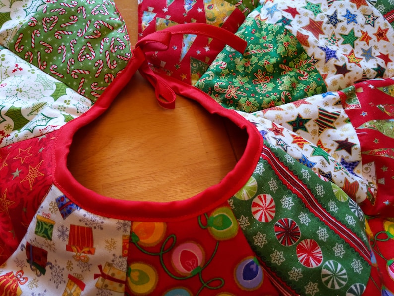 K&ouml;nnte beinhalten: Ein festlicher Weihnachtsbaumrock mit rotem Rand und einer Vielzahl von Stoffflicken mit Feiertagsmotiven. Die Flicken zeigen Zuckerstangen, Sterne, Geschenke und Lichterketten. Der Rock hat einen Durchmesser von etwa 122 cm.
