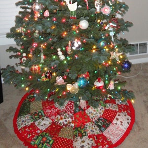 K&ouml;nnte beinhalten: Ein geschm&uuml;ckter Weihnachtsbaum mit bunten Lichtern und Ornamenten. Der Baum ist von einem Patchwork-Weihnachtsbaumrock mit roten, gr&uuml;nen und wei&szlig;en Mustern umgeben. Der Rock hat einen roten R&uuml;schenrand.
