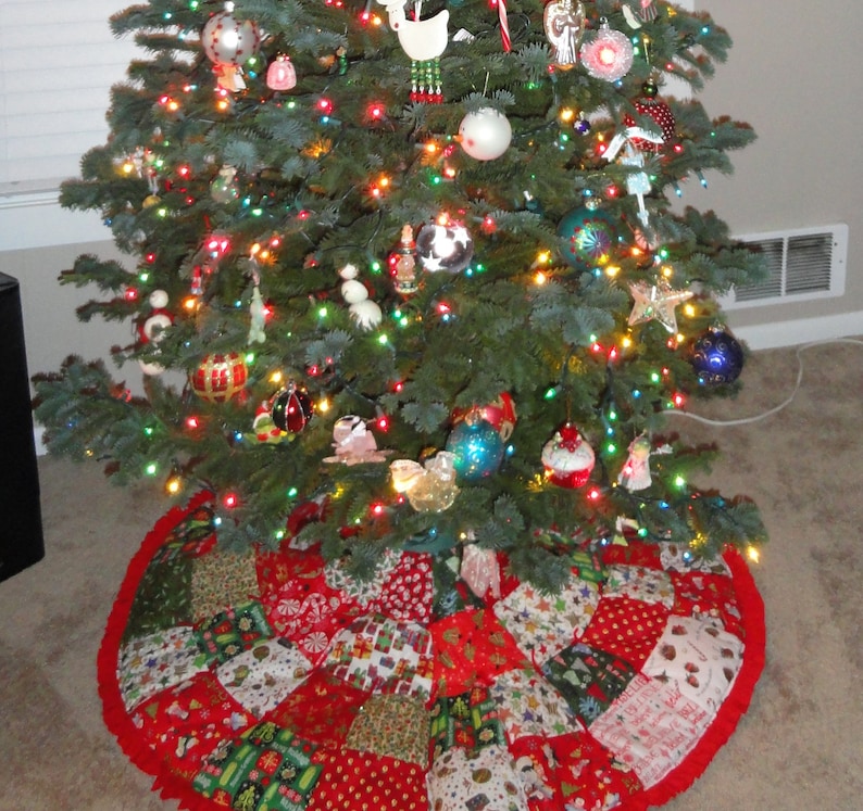 K&ouml;nnte beinhalten: Ein geschm&uuml;ckter Weihnachtsbaum mit bunten Lichtern und Ornamenten. Der Baum ist von einem Patchwork-Weihnachtsbaumrock mit roten, gr&uuml;nen und wei&szlig;en Mustern umgeben. Der Rock hat einen roten R&uuml;schenrand.
