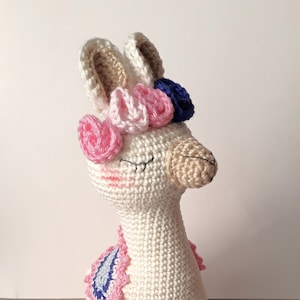 May include: A handmade white crochet llama with closed eyes, adorned with pink, blue, and white floral accents on its head. The llama has a tan snout and feet, and a decorative patch on its side. A charming, whimsical stuffed animal.