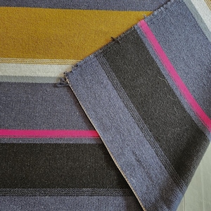 May include: A woven textile featuring a horizontal striped pattern in shades of slate blue, mustard yellow, charcoal gray, white, and a vibrant hot pink accent stripe. The fabric has a visible weave texture and a folded corner showing the reverse side.