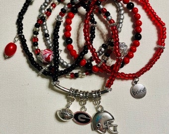 May include: A set of six beaded bracelets in red, black, and silver with charms including a football, a letter G, and a helmet. The charms are attached to a silver bar with the word "Believe" on a charm.