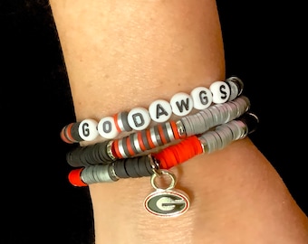 May include: A layered bracelet with black, red, and gray beads, featuring the word "GODAWGS" in white letters. A silver charm with a "G" in red is attached to the bracelet.