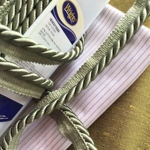 May include: A close-up of a sage green braided trim with a white label that reads "Wrights 1836321528A -3/8"-9.5MM 18 YDS/16.5M 47% COTTON 35% POLYESTER RAYON DRY CLEAN ONLY". The trim is on top of a white fabric with thin pink stripes and a green fabric.