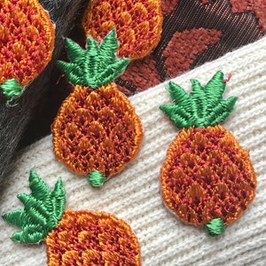 May include: Five embroidered pineapple patches in shades of orange and green. The patches are on a white and gray fuzzy surface.