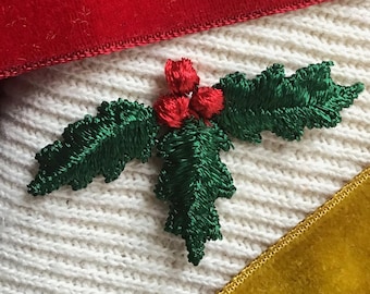 May include: Two green embroidered holly sprigs with red berries, one on a red velvet surface and the other on a white knitted surface.