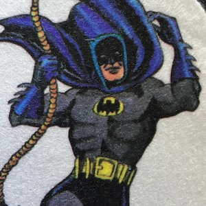 May include: A close-up of a superhero character wearing a blue cape and a black suit with a yellow belt. The character is holding a rope in their right hand and has a serious expression on their face.