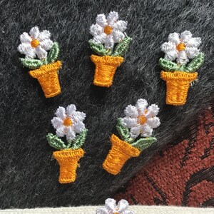 Puede incluir: Parches bordados de flores de margarita. Cada parche presenta una margarita blanca con un centro amarillo, hojas verdes y una maceta naranja. Los parches están dispuestos sobre un fondo texturizado, mostrando los detalles del diseño floral.