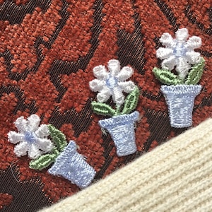 May include: Three embroidered white flowers in blue pots with green leaves on a red fabric background.