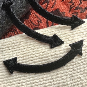 May include: Three black fabric arrows with a fuzzy texture are sewn onto a white, cream, and orange fabric background. The arrows are curved and point in different directions.