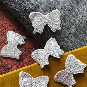 Puede incluir: Cinco adornos de lazo de lentejuelas plateadas sobre una variedad de superficies de tela. Los lazos son todos del mismo tamaño y forma, y tienen un acabado brillante e iridiscente.