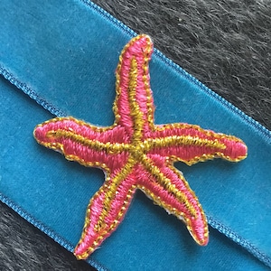 Puede incluir: Una estrella de mar bordada en rosa y dorado sobre una cinta de terciopelo azul.