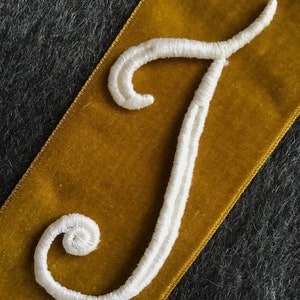 May include: A white embroidered letter "J" in a cursive style, stitched onto a gold velvet ribbon. The ribbon is set against a dark grey textured background. The embroidery uses a thick, textured thread. The image is a close-up.