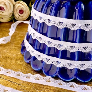 May include: White lace trim with scalloped edges, shown wrapped around a blue ceramic bowl. The lace is delicate and has a soft, feminine look.