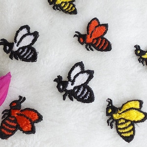 May include: Embroidered bee patches in various colors, including yellow, orange, and black, are scattered on a white, textured surface. The patches feature detailed stitching, showcasing the bee's body and wings. A pink flower and green leaves are in the lower left corner.
