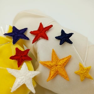 May include: Assortment of colorful star-shaped fabric patches in red, blue, yellow, and white. The patches are sewn onto a white fabric surface.