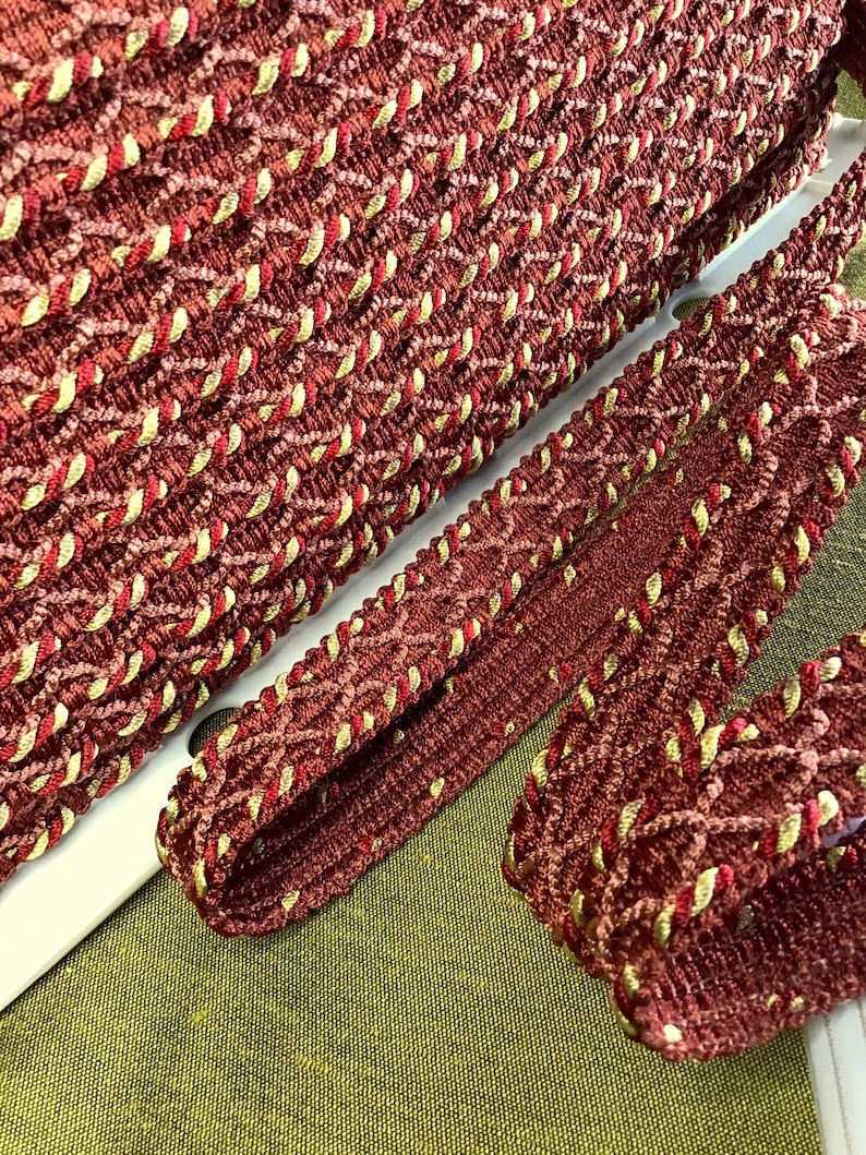 May include: Red and gold braided trim with a diamond pattern. The trim is made of a soft, textured fabric.