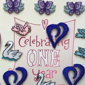 May include: A pink and white card with the text "Celebrating One Year" in pink and white lettering. The card is decorated with blue and purple butterflies, blue birds, and pink doves. There are also two blue heart-shaped designs.