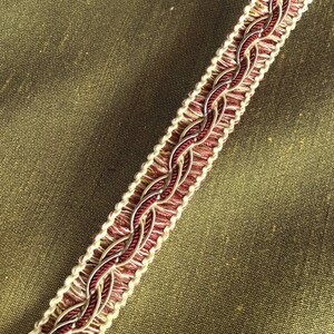 May include: A close-up of a burgundy and cream braided trim with a subtle gold thread woven throughout. The trim is on a green fabric background.