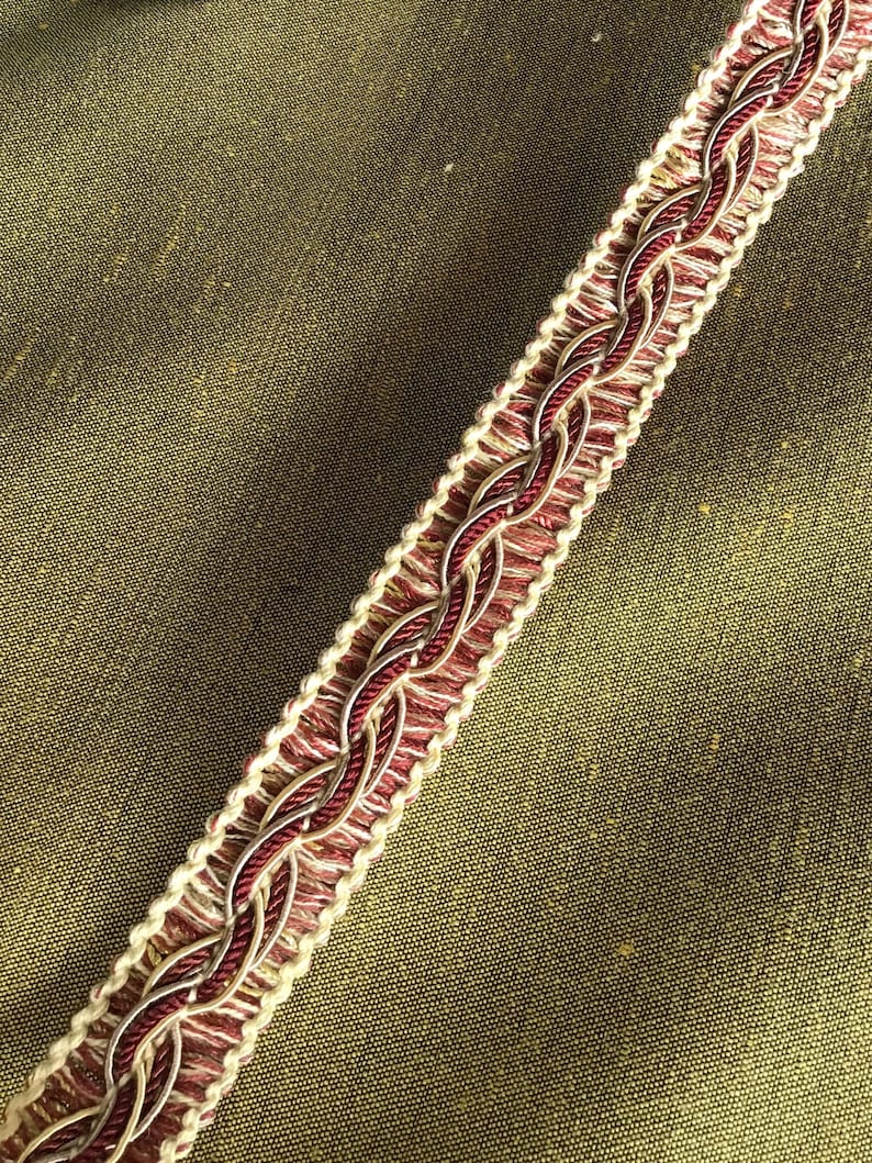 May include: A close-up of a burgundy and cream braided trim with a subtle gold thread woven throughout. The trim is on a green fabric background.