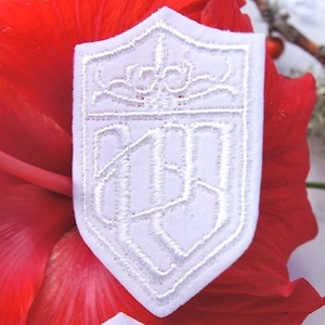 May include: White embroidered patch with a shield shape and a monogram design. The monogram is a stylized letter "H" with a crown above it.
